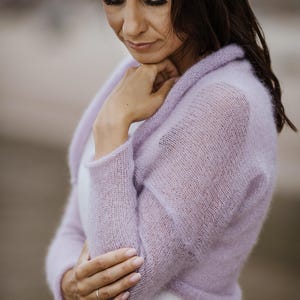 May include: A woman wearing a light purple, sheer, knitted bolero sweater. She is also wearing a white dress and a light-colored hat. The bolero has long sleeves and a rounded collar. The background is blurred.