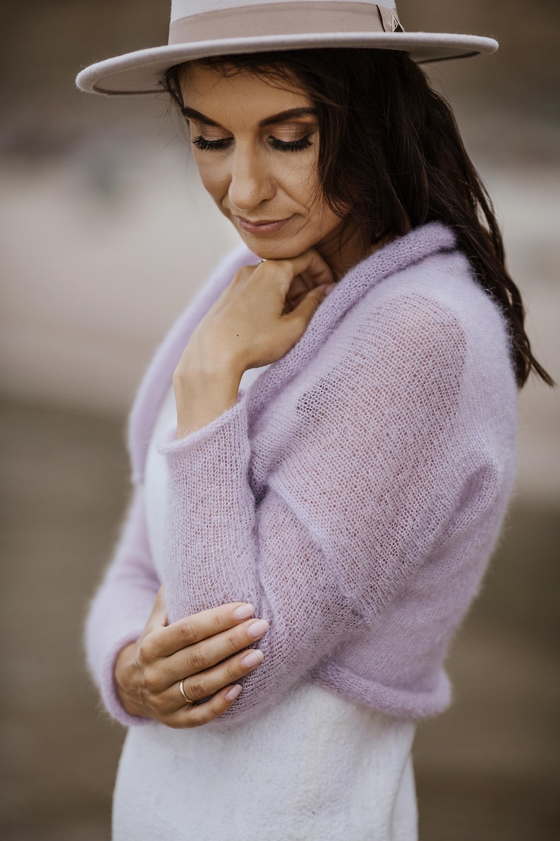 May include: A woman wearing a light purple, sheer, knitted bolero sweater. She is also wearing a white dress and a light-colored hat. The bolero has long sleeves and a rounded collar. The background is blurred.