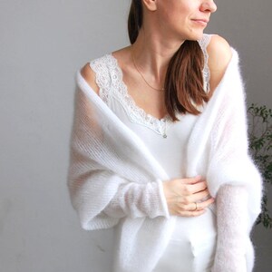 May include: A woman wearing a white, sheer, knitted shawl over a white camisole with lace trim. The outfit includes white pants. The shawl has long sleeves. The background is a light gray wall.