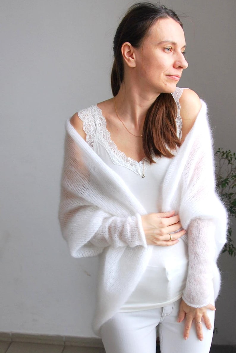 May include: A woman wearing a white, sheer, knitted shawl over a white camisole with lace trim. The outfit includes white pants. The shawl has long sleeves. The background is a light gray wall.