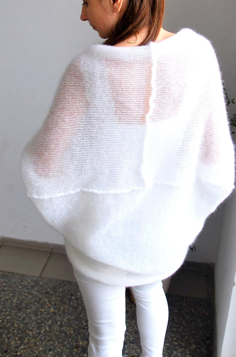 May include: A white, oversized, open-weave knit sweater. The sweater has a wide, flowing design with a layered effect. The model is wearing white pants. The sweater is made of a soft, lightweight material.