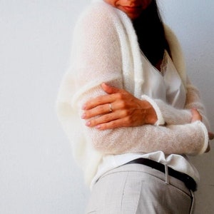 May include: Off-white, open-front cardigan with long sleeves. The cardigan is made of a sheer, lightweight knit material. The model is wearing a white camisole and light-colored plaid pants.
