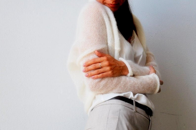 May include: Off-white, open-front cardigan with long sleeves. The cardigan is made of a sheer, lightweight knit material. The model is wearing a white camisole and light-colored plaid pants.