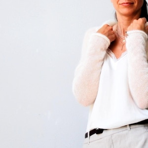 May include: A woman wearing a cream-colored, fuzzy knit sweater over a white camisole with lace trim. She is also wearing light gray pants with a black belt. A delicate necklace is visible.