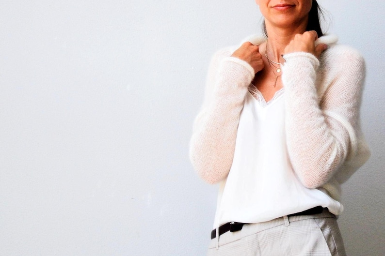 May include: A woman wearing a cream-colored, fuzzy knit sweater over a white camisole with lace trim. She is also wearing light gray pants with a black belt. A delicate necklace is visible.