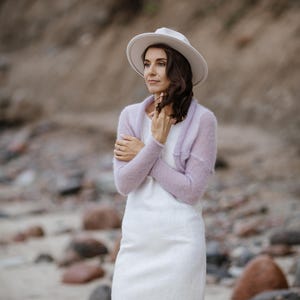 May include: A woman wearing a white dress, a light purple shrug, and a white hat. The dress is knee-length with a slight flare at the hem. The shrug is a soft knit. The hat has a wide brim.