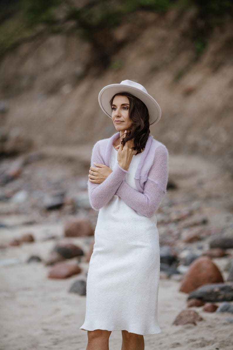 May include: A woman wearing a white dress, a light purple shrug, and a white hat. The dress is knee-length with a slight flare at the hem. The shrug is a soft knit. The hat has a wide brim.