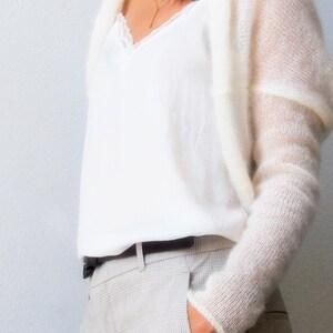 May include: A cream-colored, open-front sweater with long sleeves is worn over a white top with a lace trim. The person is wearing light gray plaid pants and a black belt. A gold necklace is visible.