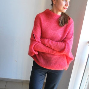 May include: A woman wearing a pink fluffy sweater and dark blue trousers. She is standing in front of a window.