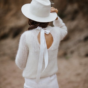 May include: A white sweater with a large bow at the back and a white hat with a small flower detail.