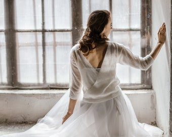 Puede incluir: Una mujer con un suéter de punto blanco con un escote en V profundo y una falda de tul blanca. Está de pie frente a una ventana con la espalda a la cámara.