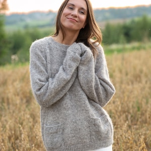 Peut inclure: Une femme portant un pull en laine bouclée brun clair avec une poche plaquée et un pantalon blanc, debout dans un champ d'herbe haute.