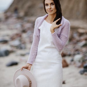 May include: A woman wearing a white dress and a light purple shrug, holding a light pink hat. The dress is knee-length with a slight flare at the hem. The hat has a brown band and a simple design. The background is a beach scene.