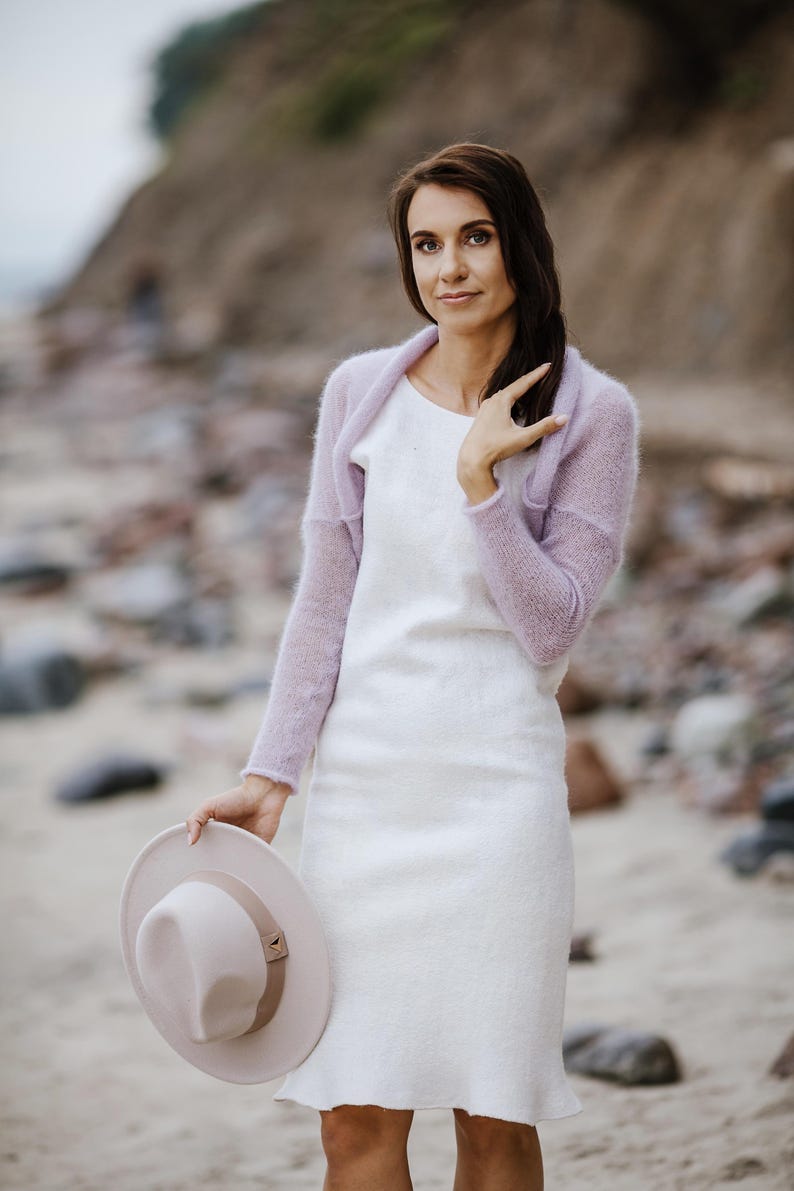 May include: A woman wearing a white dress and a light purple shrug, holding a light pink hat. The dress is knee-length with a slight flare at the hem. The hat has a brown band and a simple design. The background is a beach scene.
