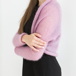May include: A light purple, open-front shrug sweater. The sweater has long sleeves and a soft, fuzzy texture. The model is wearing a black top and black pants. The background is white.