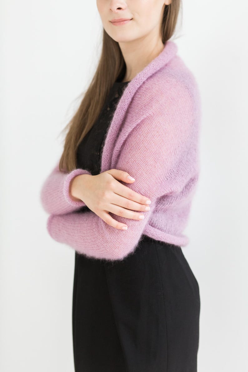 May include: A light purple, open-front shrug sweater. The sweater has long sleeves and a soft, fuzzy texture. The model is wearing a black top and black pants. The background is white.