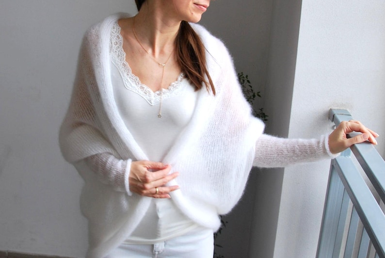 May include: A white, sheer, knitted shawl with long sleeves. The shawl is draped over a white top with lace trim. A gold necklace with a small pendant is visible.