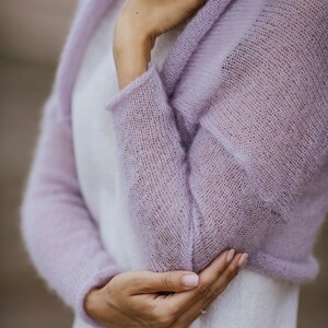 May include: A light purple, open-front knitted cardigan with long sleeves. The delicate, sheer fabric is a soft lavender hue. The cardigan is worn over a white garment, creating a layered effect. The sleeves are long and the knit has a subtle texture.