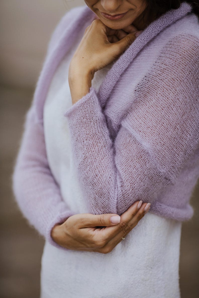 May include: A light purple, open-front knitted cardigan with long sleeves. The delicate, sheer fabric is a soft lavender hue. The cardigan is worn over a white garment, creating a layered effect. The sleeves are long and the knit has a subtle texture.