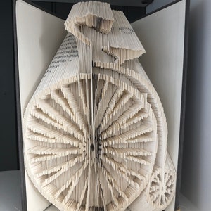 Könnte beinhalten: Eine Buchkunstskulptur eines Vintage-Fahrrads, hergestellt aus den Seiten eines Buches. Die Seiten sind gefaltet und übereinander gelegt, um einen 3D-Effekt zu erzeugen. Die Skulptur wird auf einer weißen Oberfläche ausgestellt.