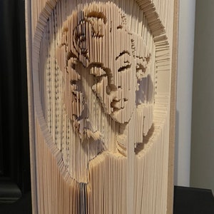 Può includere: Una scultura di arte libraria di un viso creata piegando le pagine di un libro. La scultura è realizzata con un libro con una copertina di colore crema.