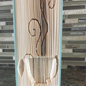 May include: A book sculpture featuring a coffee cup design carved into the pages. Steam rises from the cup, created by the folded pages. The book has a pale blue cover and is displayed against a neutral backdrop.
