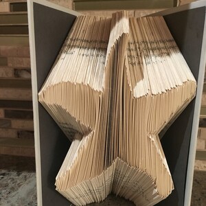 May include: A book folded into the shape of a star, displayed on a gray stand. The book is made of old, yellowed pages and is held together with a brown string.
