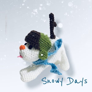 Puede incluir: Adorno de perro de ganchillo con un sombrero marrón y verde, una bufanda azul y una pequeña nariz naranja. El perro tiene una pequeña lengua rosa que sobresale. El texto "Snowy Days" está escrito en azul en cursiva.