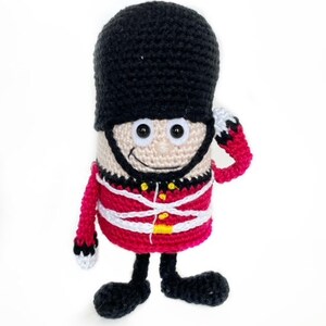 May include: A crocheted toy soldier wearing a red and white uniform with a black hat. The toy has a smiling face with black eyes and a black mustache.