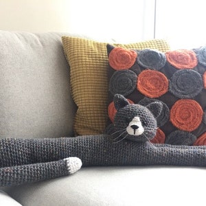 May include: A handmade, grey crochet cat-shaped cushion with white accents, stretched out on a grey sofa. The cat has a white muzzle and whiskers. A decorative cushion with orange and grey circles is behind it.
