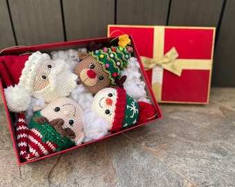 May include: A red gift box with a white bow is open, revealing five crocheted Christmas ornaments. The ornaments are a snowman, a reindeer, Santa Claus, an elf, and a Christmas tree. The ornaments are all white, red, and green.