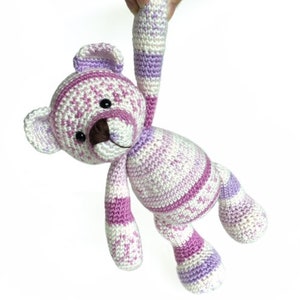 May include: A crocheted teddy bear with pink and white stripes. The bear has a brown nose and black eyes.
