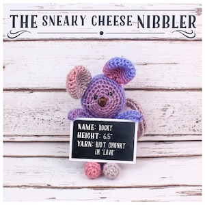 Könnte beinhalten: Eine gehäkelte Maus hält ein Schild mit der Aufschrift "NAME: ROCKY, HEIGHT: 6.5", YARN: RIOT CHUNKY IN 'LAVA'. Die Maus ist lila und rosa und sitzt auf einem weißen Holzgrund.