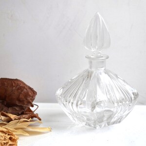 May include: A clear glass bottle with a faceted design and a pointed stopper. The bottle is empty and sits on a white surface.