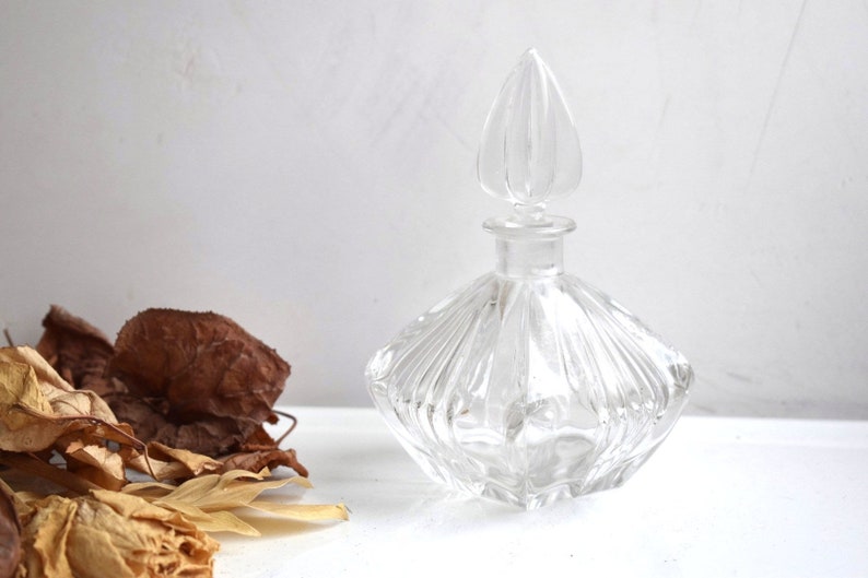 May include: A clear glass bottle with a faceted design and a pointed stopper. The bottle is empty and sits on a white surface.