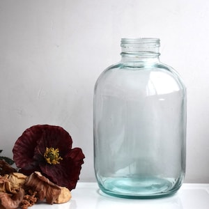 Könnte beinhalten: Eine klare Glasflasche mit einer breiten Öffnung und einem abgerundeten Körper. Die Flasche steht auf einer weißen Oberfläche.