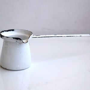 Puede incluir: Cafetera de esmalte blanco con borde y asa negros. La cafetera tiene un pequeño pico y está diseñada para preparar café turco.