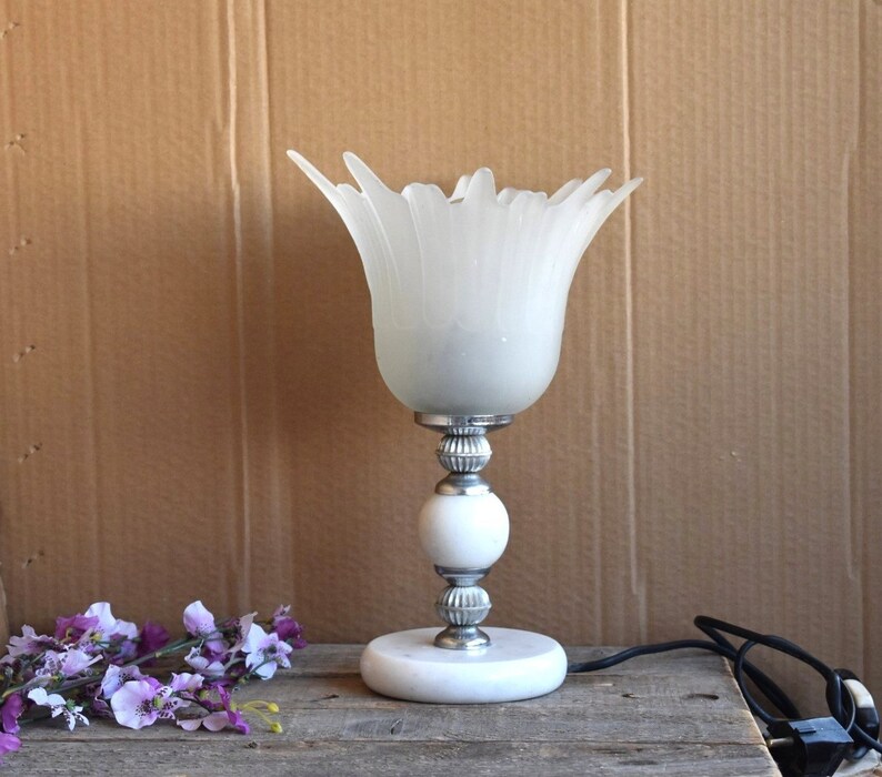 May include: White frosted glass table lamp with a flower-shaped shade and a white marble base. The lamp has a silver metal stem and a silver metal base.