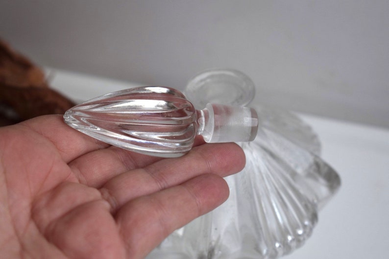 May include: A clear glass bottle with a decorative, fluted stopper. The bottle has a scalloped design and is perfect for holding perfume or essential oils.