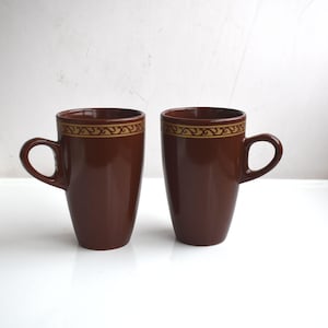 Può includere: Due tazze da caffè in ceramica marrone con una fascia decorativa dorata vicino al bordo. Le tazze hanno un design semplice ed elegante con un manico curvo. Le tazze sono poste una accanto all'altra su una superficie bianca.
