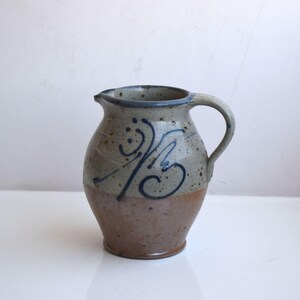 Può includere: Un piccolo boccale in ceramica fatto a mano con una glassa marrone, bianca e blu. Il boccale ha una forma semplice e arrotondata e un'unica maniglia. Presenta un design astratto blu e bianco.