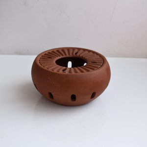 May include: A terracotta-colored ceramic tea warmer. The round warmer has a central opening and a ridged top. Small, evenly spaced holes are around the base of the warmer. The tea warmer is on a white surface.