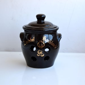 Puede incluir: Un tarro de ajo de cerámica negra con tapa y dos asas. El tarro tiene agujeros de ventilación y letras doradas que dicen "Zwiebel Knoblauch" con una ilustración de un bulbo de ajo.