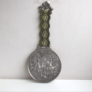 May include: A silver-colored, round decorative wall hanging with a detailed relief scene. It features a green and gold woven strap and an ornate top. The relief depicts figures on horseback and other detailed elements.