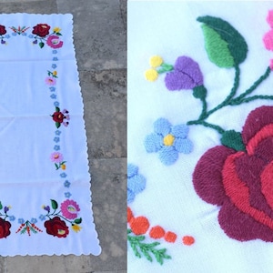 Könnte beinhalten: Weißer rechteckiger Tischläufer mit gewelltem Rand, bestickt mit bunten Blumen. Das florale Design umfasst rote, rosa, blaue und gelbe Blüten und schafft ein lebendiges und dekoratives Stück.