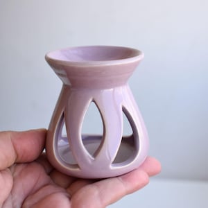 Op de afbeelding: Een lavendelkleurige keramische etherische olie diffuser met een afgeronde basis en een uitlopende bovenkant. De diffuser heeft drie grote, druppelvormige uitsparingen. De bovenkant heeft een kleine kom voor het vasthouden van olie en water. Een decoratief item voor aromatherapie.