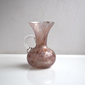 Może przedstawiać: Mały, przezroczysty szklany wazon z różowo-białym wzorem w spiralę i pojedynczym uchwytem. Wazon ma okrągłe dno i wąską szyjkę.