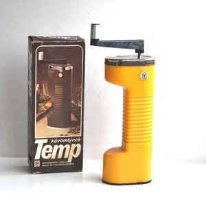Op de afbeelding: Gele handmatige koffiemolen met een zwarte handgreep en een bruine doos met de tekst "kavómlýnek Temp" erop. De doos vermeldt ook "Výrobný závod Semily Made in Czechoslovakia".
