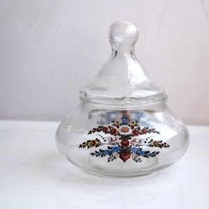 K&ouml;nnte beinhalten: Ein klares Glasgef&auml;&szlig; mit Deckel, verziert mit einem Blumendesign in Rot-, Blau-, Gelb- und Gr&uuml;nt&ouml;nen. Das Gef&auml;&szlig; hat einen runden K&ouml;rper und einen gew&ouml;lbten Deckel.