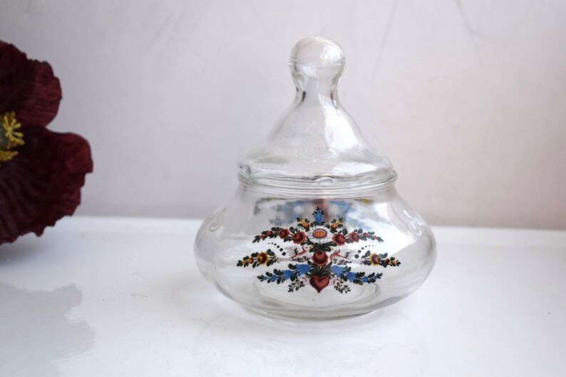 K&ouml;nnte beinhalten: Ein klares Glasgef&auml;&szlig; mit Deckel, verziert mit einem Blumendesign in Rot-, Blau-, Gelb- und Gr&uuml;nt&ouml;nen. Das Gef&auml;&szlig; hat einen runden K&ouml;rper und einen gew&ouml;lbten Deckel.